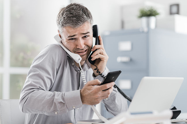 Image of young man on 2 landline handsets and smartphone looking stressed